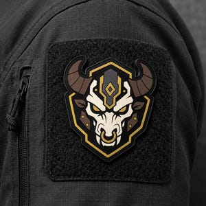 May include: A black tactical patch featuring a stylised bull head design with brown horns, a cream-coloured face, and gold accents, attached to a black ripstop fabric sleeve with a zippered pocket and a hook-and-loop fastener backing.