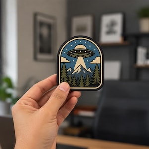 May include: A die-cut sticker featuring a graphic illustration of a flying saucer hovering over a mountain range and a forest of pine trees. The design includes a blue sky with white clouds, stars, and a bright light beam shining down.