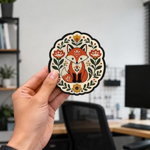 May include: A circular sticker featuring a stylized orange fox sitting in the center, surrounded by a symmetrical pattern of dark green leaves, stems, and orange and yellow flowers on a cream-colored background with a thick black border.