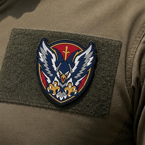 May include: A tactical morale patch featuring a stylized eagle with spread wings, sharp talons, and a yellow beak. The design includes a red circular background with a gold cross, set against a dark olive green fabric base with a hook backing.