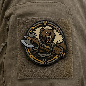 May include: A circular morale patch featuring a brown bear with an open mouth holding a silver battle axe. The bear is set against a cream-colored fur background within a black, gold, and grey circular frame, attached to olive drab fabric.