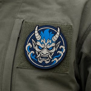 May include: A circular patch featuring a stylised blue and white Oni mask with horns, sharp teeth, and glowing blue eyes, surrounded by blue wave patterns. The patch is attached to an olive green fabric surface with a visible vertical zipper.