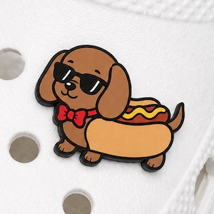 May include: A shoe charm featuring a brown dachshund dog wearing black sunglasses and a red bow tie. The dog is depicted inside a tan hot dog bun with a brown sausage and a yellow line of mustard on its back, attached to a white textured shoe.