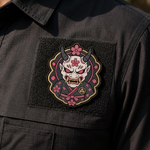May include: A circular patch featuring a white oni mask with horns, red eyes, and gold fangs, surrounded by pink cherry blossoms and a dark gray border. The patch is attached to a black fabric shirt with a textured grid pattern and a square velcro base.