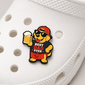 Può includere: Un charm per scarpe raffigurante un cane giallo con un berretto rosso, occhiali da sole neri e un grembiule nero con la scritta BEST DAD EVER e l'icona di una spatola e una forchetta. Il cane tiene in mano un boccale di birra su una scarpa.
