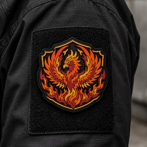 Puede incluir: Un parche rectangular negro con un emblema central en forma de escudo que muestra un fénix estilizado surgiendo de las llamas. El fénix está representado en tonos vibrantes de naranja, rojo y amarillo sobre un fondo oscuro en una manga negra.