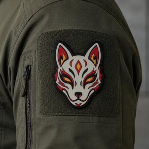 May include: An olive green tactical jacket sleeve featuring a square hook-and-loop patch area. Attached is a rubberized patch displaying a stylized white fox mask with red, black, and yellow accents, featuring pointed ears and sharp facial markings.