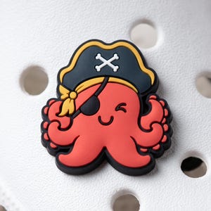 May include: A red octopus shoe charm featuring a black pirate hat with a white skull and crossbones emblem. The octopus wears a black eye patch over one eye, has a smiling mouth, and is positioned on a white surface with circular cutouts.