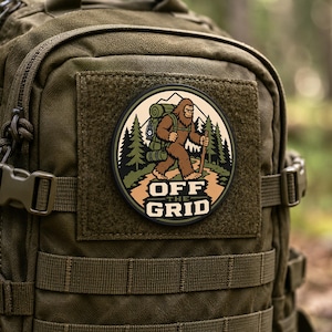 Op de afbeelding: Een ronde geborduurde patch bevestigd op een olijfgroene tactische rugzak. De patch toont een bruin Bigfoot-figuur met een groene rugzak en wandelstok in een bosrijke omgeving met de tekst OFF THE GRID in dikgedrukte witte letters.