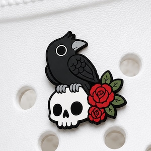 Könnte beinhalten: Ein Schuh-Anstecker mit einem schwarzen Raben, der auf einem weißen menschlichen Schädel sitzt, begleitet von zwei roten Rosen mit grünen Blättern. Der Anstecker ist an einem weißen perforierten Schuh befestigt und zeigt ein grafisches Design.
