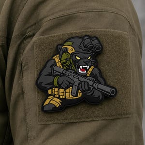 Pode incluir: Um emblema tático com uma pantera preta usando um capacete escuro com óculos de visão noturna e um colete tático amarelo. A pantera segura um rifle preto com silenciador. O emblema está fixado em um tecido de cor verde-oliva.
