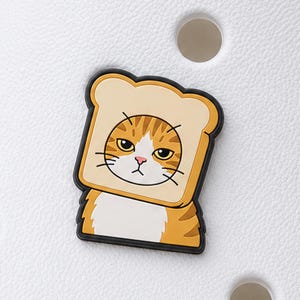 May include: A shoe charm featuring a cartoon orange tabby cat with a white chest, wearing a slice of bread around its face. The cat has yellow eyes, a pink nose, and a serious expression, outlined in black against a white textured background.