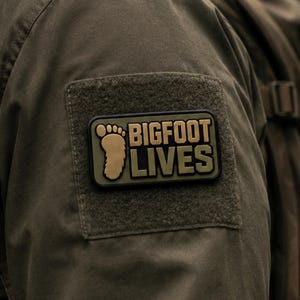 May include: A rectangular olive green patch with a black border featuring a tan footprint icon and the text BIGFOOT LIVES in bold tan lettering. The patch is attached to a dark olive green fabric surface with a matching square hook and loop panel.