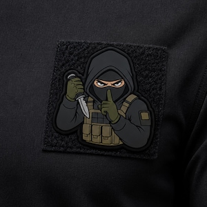 May include: A square black tactical patch featuring a cartoon illustration of a hooded figure in a black balaclava and dark clothing. The figure wears a tan tactical vest, olive green gloves, and holds a silver knife while making a hush gesture.