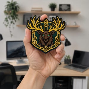 May include: A shield-shaped patch featuring a brown deer head with large yellow antlers, set against a dark green background. The design includes a yellow border with geometric patterns and a small yellow Valknut symbol at the bottom center.