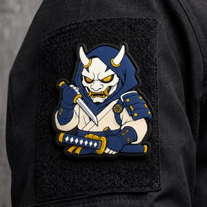 Peut inclure: Un écusson rectangulaire noir à scratch représentant un personnage de dessin animé portant une capuche bleue et un masque blanc à cornes. Le personnage tient une petite lame et un katana, vêtu d'une armure aux accents bleus et blancs.
