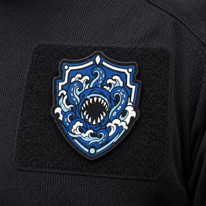 May include: A black tactical patch featuring a shield-shaped design with a blue and white graphic of a sea monster with tentacles and sharp teeth emerging from ocean waves. The patch is attached to a black textured fabric surface with hook backing.