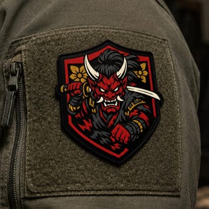 Peut inclure: Un écusson en forme de bouclier représentant un démon oni au visage rouge avec des cornes blanches et des dents pointues, tenant un katana. Le motif inclut une armure noire et rouge, des détails floraux dorés sur un fond vert olive.