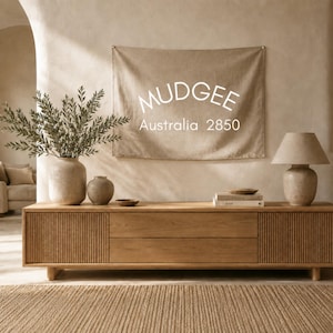 May include: A rectangular beige fabric wall hanging featuring the white text MUDGEE Australia 2850. The banner is displayed above a long wooden media console with vertical slat detailing, two vases with greenery, and a table lamp with a beige shade.