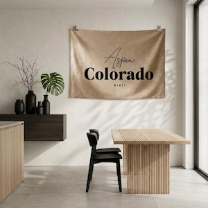Könnte beinhalten: Ein rechteckiger Wandbehang aus beigem Stoff mit dem schwarzen Text Aspen, Colorado, und der Zahl 81611. Der Wandbehang ist mit zwei silbernen Metallclips an einer hellen Wand befestigt und befindet sich über einem Holztisch und Stuhl.