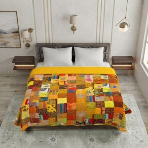Puede incluir: Una colcha rectangular con un diseño de patchwork de varios cuadrados de tela en tonos amarillo, naranja, rojo y marrón. La colcha cubre una cama con dos almohadas blancas, situada entre dos mesitas de noche de madera y lámparas de pared.