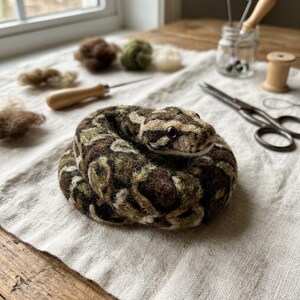 May include: A needle-felted snake figurine coiled in a circle, featuring a textured pattern of dark brown, olive green, and cream-colored wool fibers. The snake has a smooth, dark, round eye and a detailed, realistic head shape crafted from soft wool.