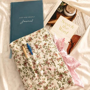 May include: A blue journal with the text Love God Greatly Journal, a book titled Search the Word, and a cream-colored fabric cover featuring a floral pattern of small pink roses and green leaves, with two pens tucked into a front pocket.