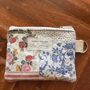 May include: A rectangular fabric zipper pouch featuring a patchwork design of floral patterns in pink, yellow, and blue on a white background. A white label is stitched to the center with the text "HANDMADE WITH LOVE Amy Contreras".