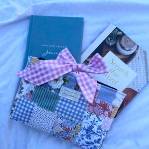 May include: A rectangular fabric pouch featuring a patchwork design of various floral and gingham patterns in blue, white, and pink tones. A large pink and white gingham bow is attached to the top. A small rectangular label reads Handmade with love.