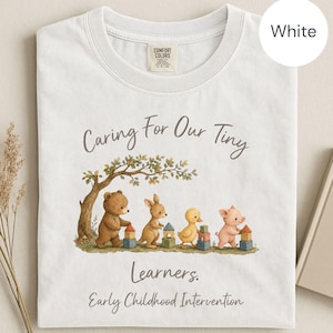 May include: A white short-sleeved t-shirt featuring a graphic of a bear, kangaroo, duck, and pig playing with wooden blocks under a tree. The text reads Caring For Our Tiny Learners. Early Childhood Intervention. The shirt has a Comfort Colors label.