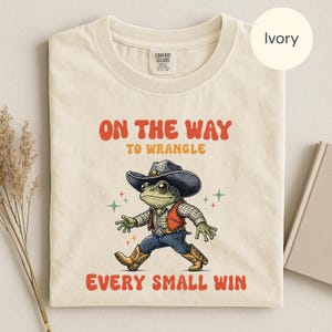 Könnte beinhalten: Ein elfenbeinfarbenes T-Shirt mit einer Grafik eines grünen Frosches, der einen Cowboyhut, ein kariertes Hemd, eine Weste, eine blaue Jeans und Stiefel trägt. Der Text ON THE WAY TO WRANGLE EVERY SMALL WIN steht in orange und gelb.