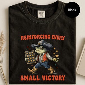 May include: A black t-shirt featuring a graphic of a frog wearing a cowboy hat, vest, and boots. The frog holds a board with twelve stars and is surrounded by small star icons. The text reads REINFORCING EVERY SMALL VICTORY in orange letters.