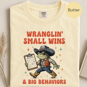 May include: A butter-colored t-shirt featuring a graphic of a frog wearing a cowboy hat and vest, holding a clipboard labeled Data Collection. The text reads Wranglin' Small Wins and Big Behaviors, surrounded by small colorful star icons.