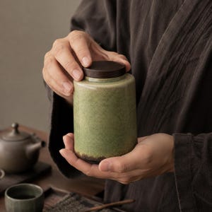 May include: A cylindrical ceramic tea canister with a mottled light green glaze and a dark brown base. The container features a flat, circular dark brown wooden lid that sits flush on the top rim, creating a simple and functional storage vessel.
