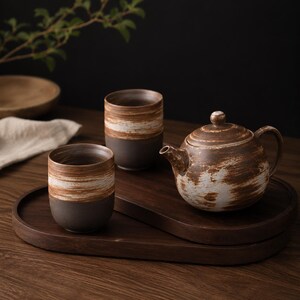 May include: A ceramic tea set consisting of a round teapot and two cylindrical cups, all featuring a dark brown base with a textured, horizontal white and light brown brushstroke pattern, arranged on a dark wooden oval serving tray.