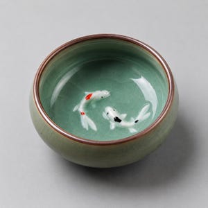Puede incluir: Un cuenco de té redondo de cerámica con un acabado de esmalte craquelado verde claro. El interior presenta dos pequeñas figuras de peces koi en relieve, una con marcas rojas y blancas y la otra con marcas negras y blancas, en el fondo.