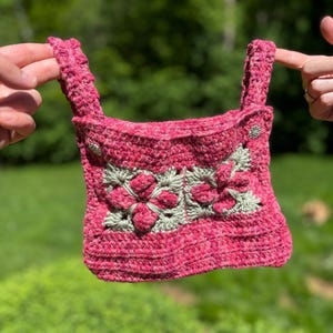 May include: A handmade crochet top featuring a vibrant pink yarn base with two prominent square motifs in sage green and pink. The garment includes two thick shoulder straps and two small, circular, silver-toned metallic buttons at the top corners.