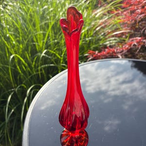 May include: A tall, vibrant red glass bud vase featuring a slender, elongated neck that flares into a ruffled, petal-like opening at the top. The base is circular and weighted, while the body displays vertical ribbed detailing throughout the glass.