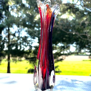May include: A tall, slender glass bud vase featuring a flared, ruffled rim. The body of the vase is crafted from clear glass with vertical streaks of deep red and dark blue pigment running from the base up toward the opening, creating a vibrant, multi-colored effect.