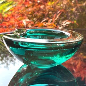 May include: A thick, translucent teal glass ashtray with a smooth, rounded bowl shape and a single indented notch on the rim. The glass features a polished finish that catches the light, creating a clear, vibrant green-blue hue throughout the piece.