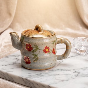 Può includere: Una teiera in ceramica grigio chiaro maculata con coperchio, decorata con fiori rossi dipinti a mano, foglie verdi e dettagli dorati. Accanto alla teiera ci sono due piccole tazze in vetro sfaccettato e un anello con perla bianca.