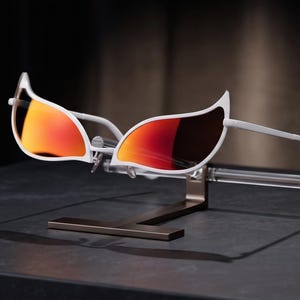 May include: Sunglasses with white frames and distinctively pointed, wing-like upper corners. The lenses feature a reflective, gradient orange and red tint. The glasses are displayed on a clear horizontal support bar attached to a dark metal stand.