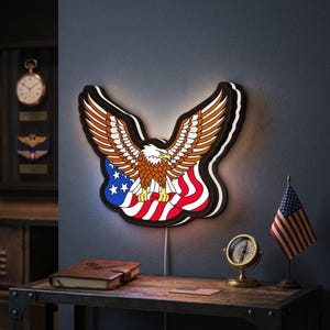 Puede incluir: Un aplique de pared LED con forma de águila calva con las alas extendidas, posicionado sobre un diseño estilizado de la bandera estadounidense en colores rojo, blanco y azul. El águila tiene plumas marrones, cabeza blanca y garras amarillas.