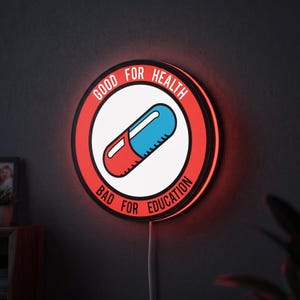 May include: A circular LED wall sign featuring a red border with the white text "GOOD FOR HEALTH" and "BAD FOR EDUCATION" surrounding a central graphic of a red and blue pill on a white background, illuminated with a soft red glow.