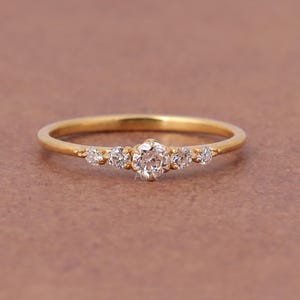 Peut inclure: Une fine bague en or avec une pierre précieuse ronde et transparente au centre, entourée de deux pierres plus petites de chaque côté, totalisant cinq pierres serties horizontalement sur l'anneau en or jaune poli.