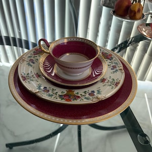 May include: A porcelain tea cup and saucer set featuring a deep magenta rim with gold trim, resting on a larger cream-colored plate decorated with a colorful floral pattern and a central urn motif, all placed atop a large, textured magenta charger.