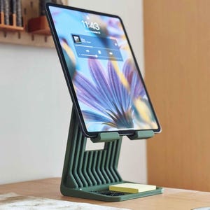 May include: A green tablet stand with a built-in tray holding a yellow sticky note pad. The tablet displays a digital interface with a floral background. The stand is on a wooden surface.