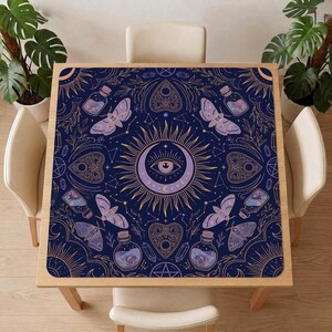 Può includere: Una tovaglia quadrata con sfondo blu scuro e un motivo ripetuto di falene, bottiglie di vetro, cristalli, soli, lune e forme di planchette in linee dorate e viola chiaro. Il design centrale mostra un grande occhio sole e luna.