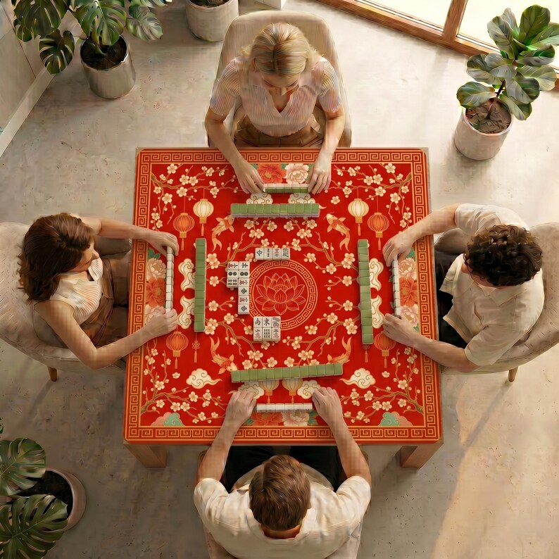 Overhead view of four people playing mahjong on a table covered with the red lantern koi lotus mahjong mat, surrounded by plants and natural light.