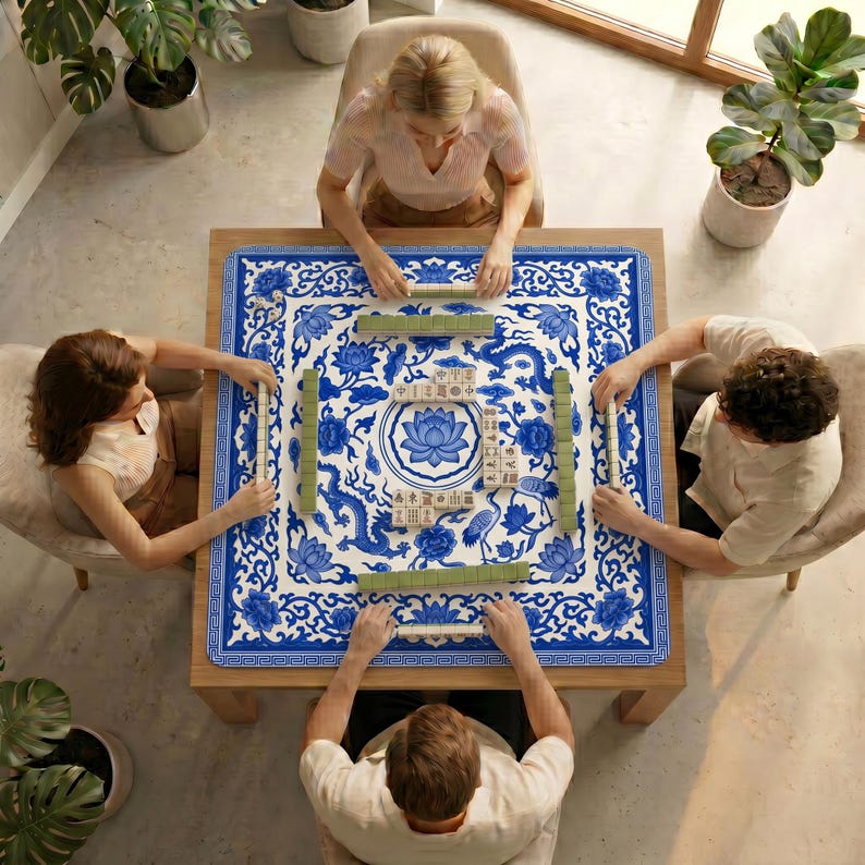 Overhead view of four people playing mahjong on a table covered with the blue and white dragon lotus mahjong mat, surrounded by plants and natural light.
